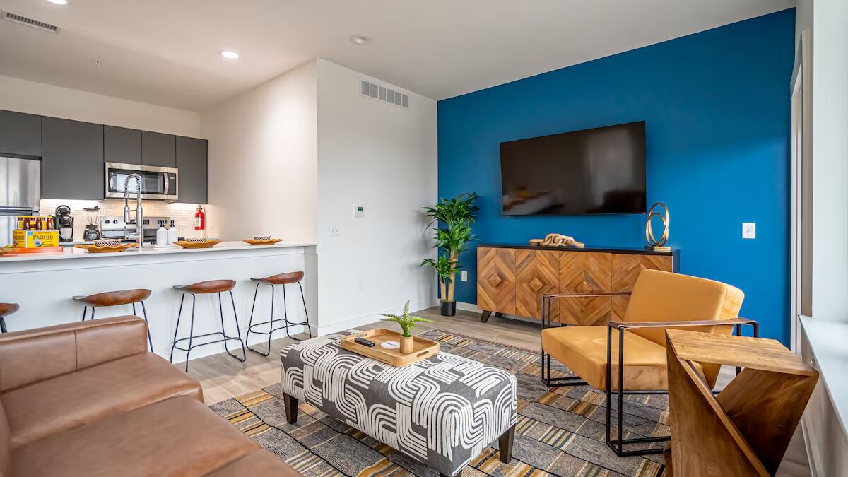 A modern living room with a sofa, ottoman, and a coffee table. There is a kitchen area with a microwave and bar stools. A TV is mounted on a blue wall.