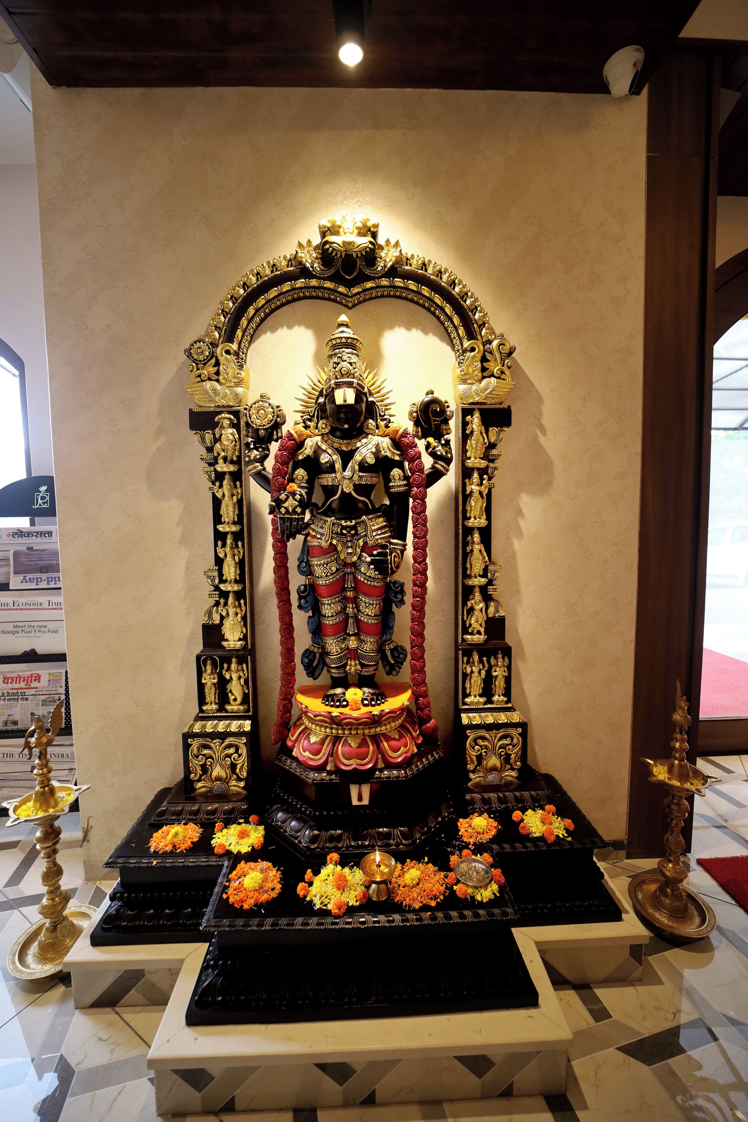 Une statue d’une divinité, posée sur un piédestal noir orné de motifs dorés, entourée de fleurs et de brûleurs d’encens.