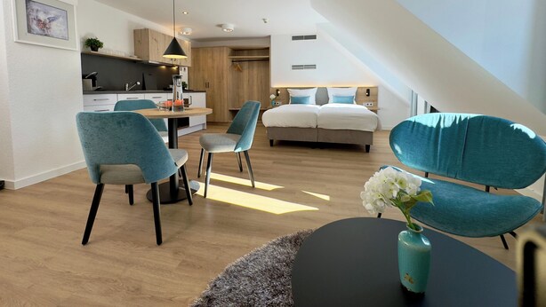 Ein modernes, helles Schlafzimmer mit einem großen Bett, einem Essbereich mit Stühlen und einer Küche im Hintergrund.
