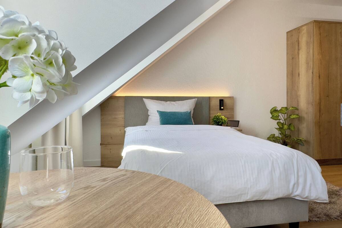 Een moderne slaapkamer met een schuine plafond, een houten hoofdbord, een wit bed met een blauwgroen kussen, een nachtkastje met een glas, en een potplant.
