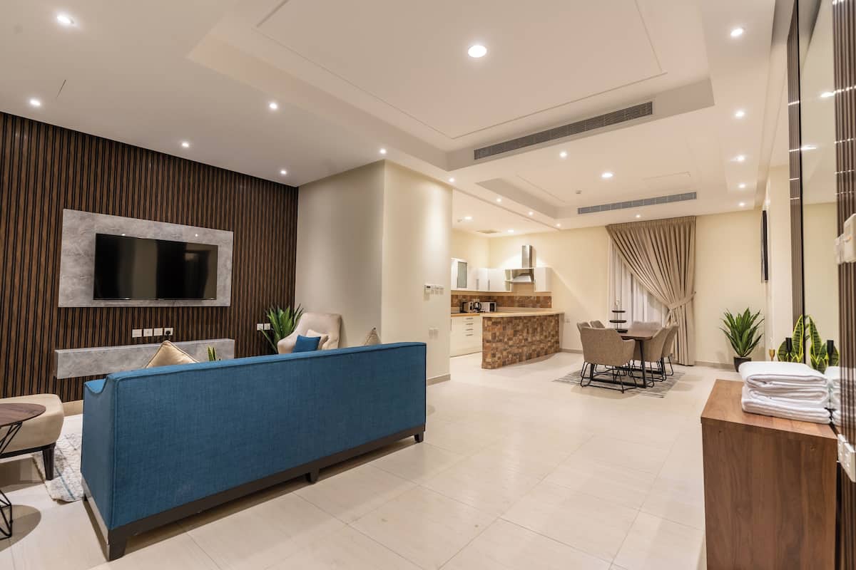 A modern living room with a blue sofa, a flat-screen TV mounted on a wooden panel, a kitchen area with a bar, and a dining set.