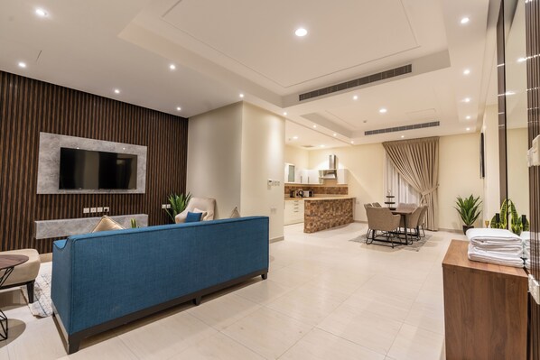 A modern living room with a blue sofa, a flat-screen TV mounted on a wooden panel, a kitchen area with a bar, and a dining set.