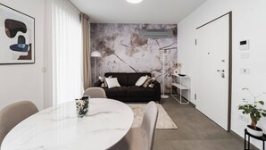 Ein modernes Wohnzimmer mit einem runden Marmortisch, einer dunkelgrauen Couch mit Kissen, einer Stehlampe und einem an der Wand befestigten Regal mit Dekorationselementen.