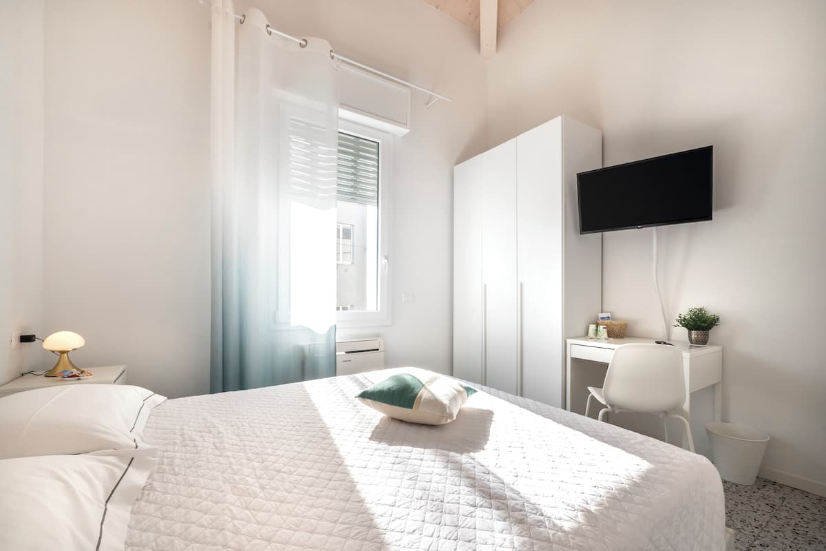 A bedroom with a bed, a desk, a chair, a TV, and a window with curtains.