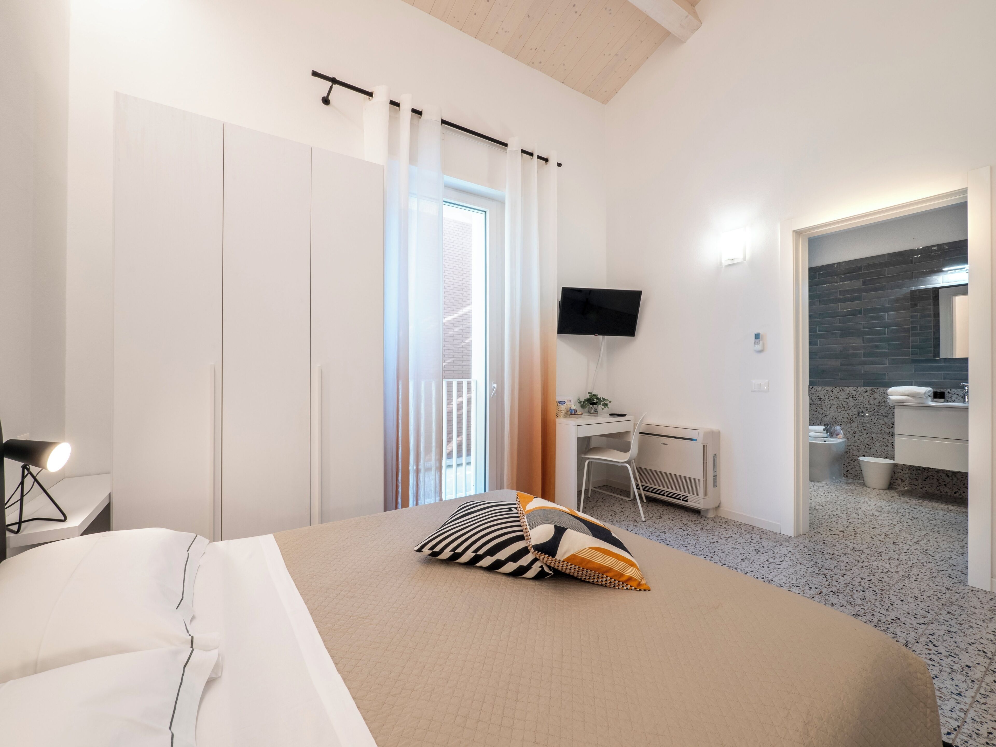A modern hotel room with a bed, a desk, a TV, and a bathroom visible through an open door.