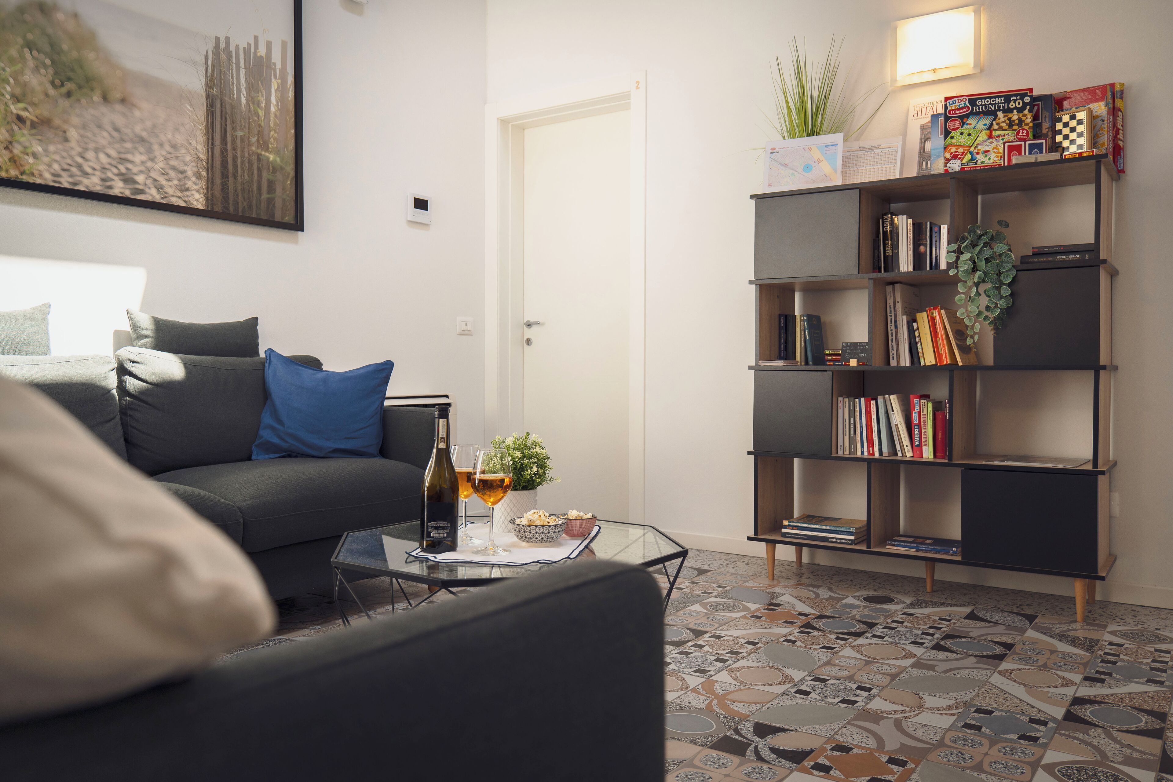 A modern living room with a grey sofa, a glass coffee table with a bottle and glasses, a bookshelf with books and decorative items, and a patterned floor.