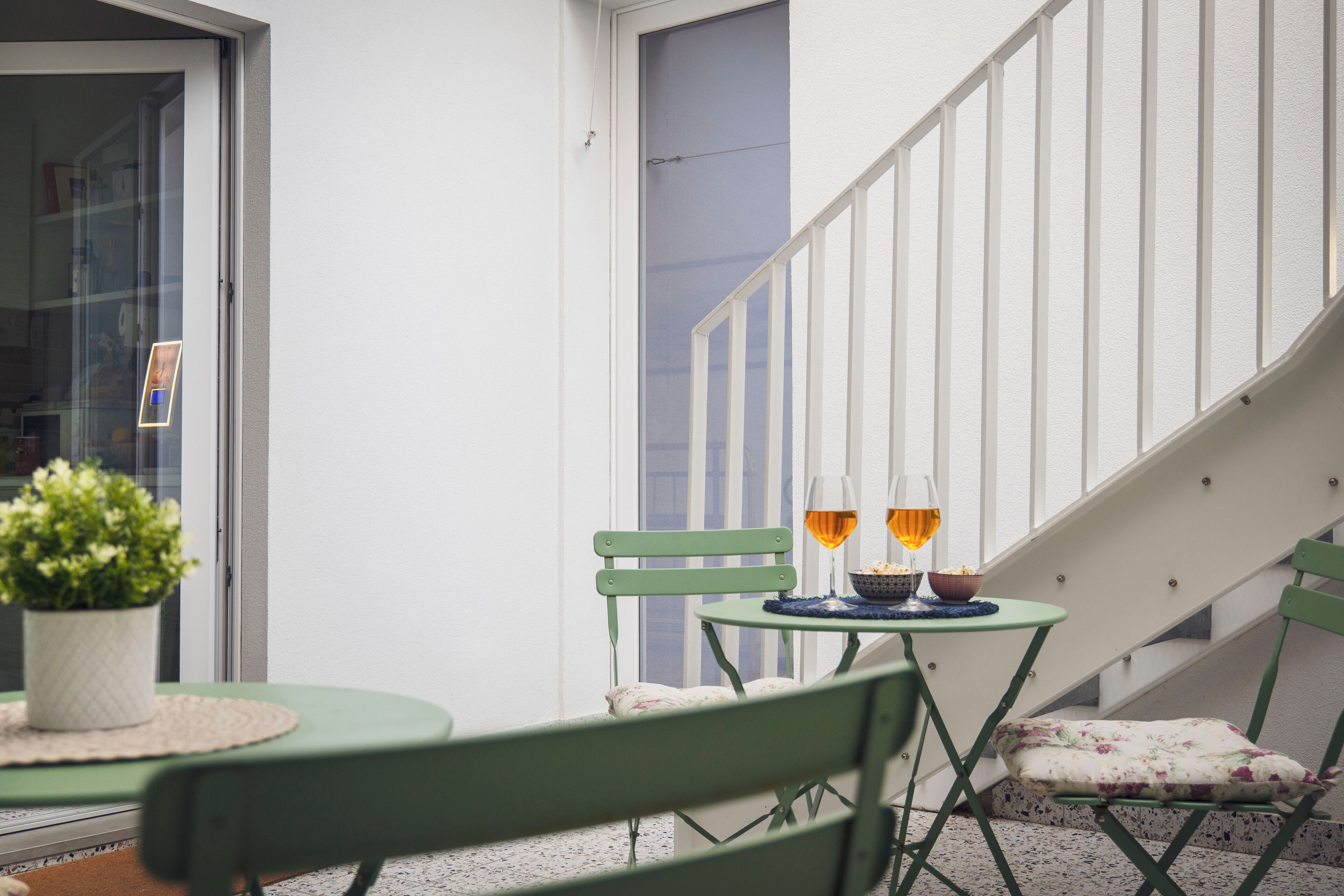A balcony with a green chair, a small table with a bowl and wine glasses, and a potted plant.