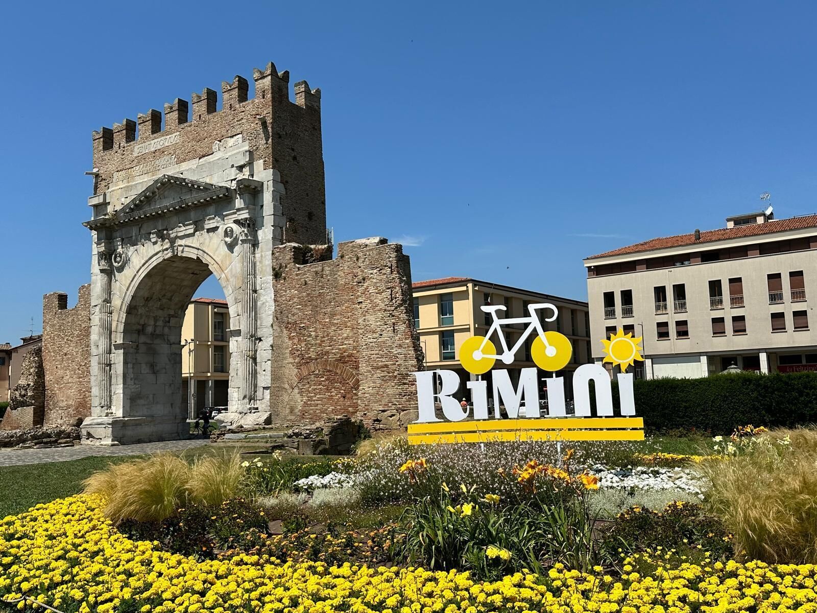 An archway with a sign reading "Rimini" and a bicycle symbol, surrounded by yellow flowers.