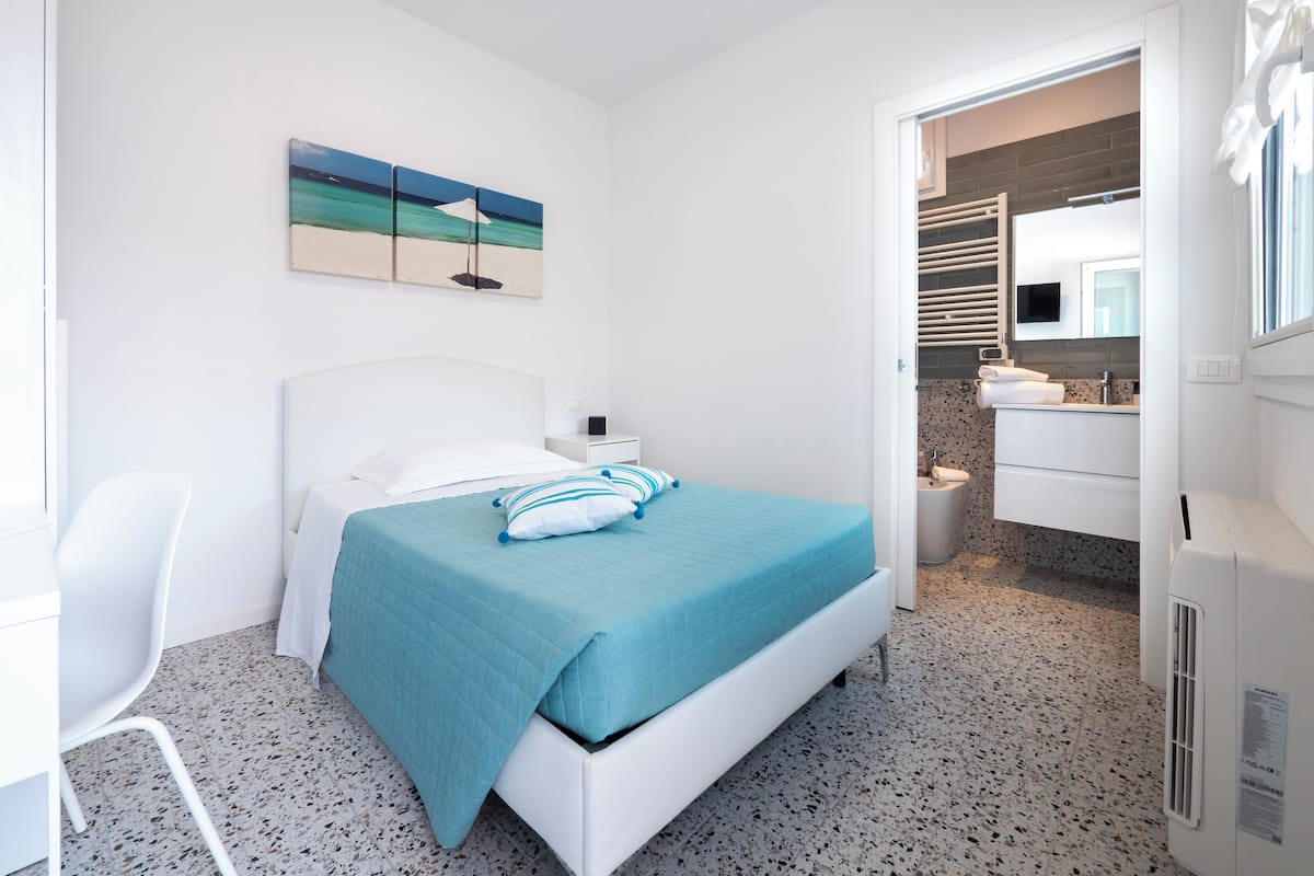 A bedroom with a bed, a nightstand, a chair, a painting, and a bathroom visible through an open door.