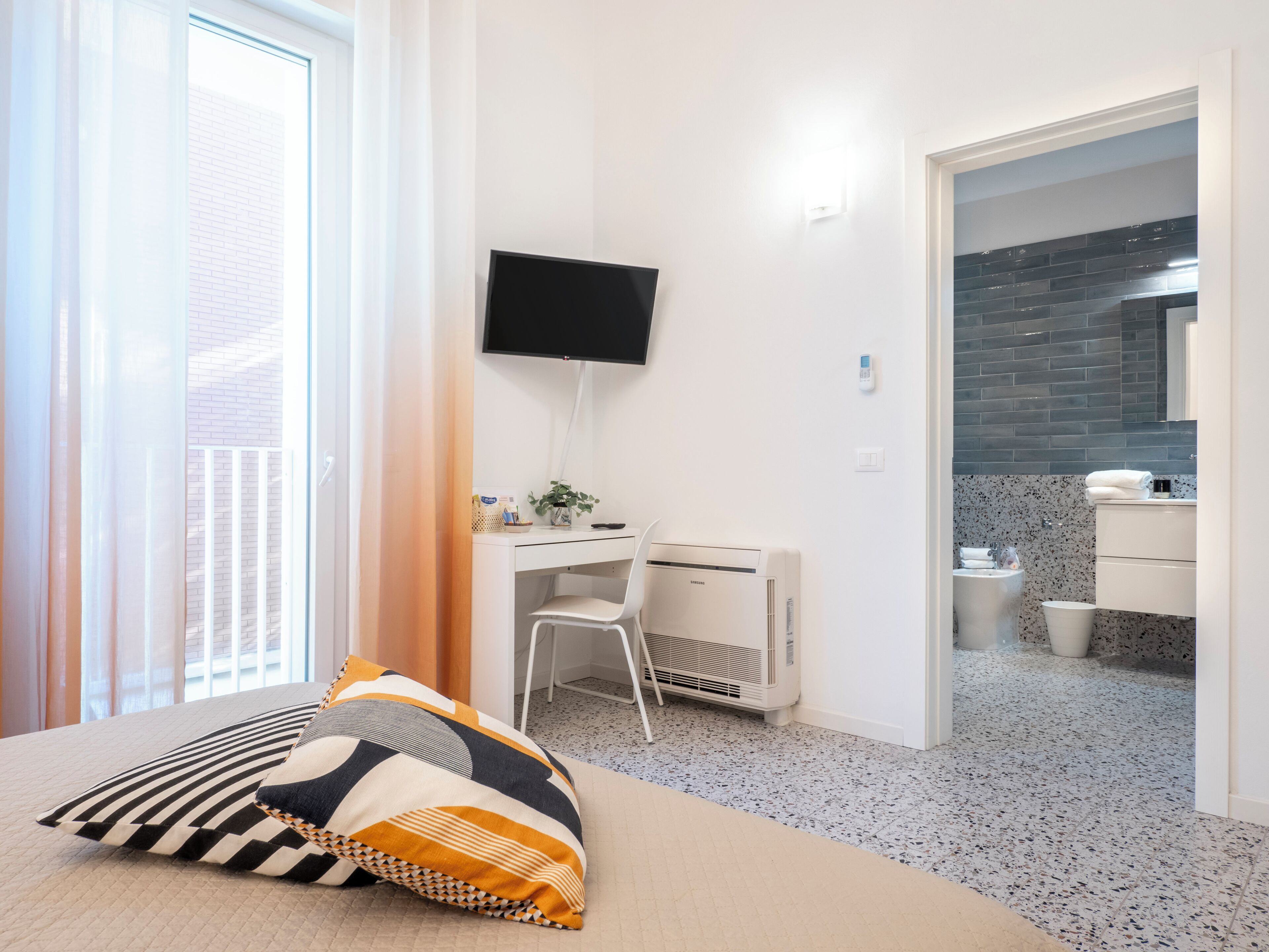 A modern hotel room with a bed, a desk with a chair, a TV, and a bathroom visible through an open door.