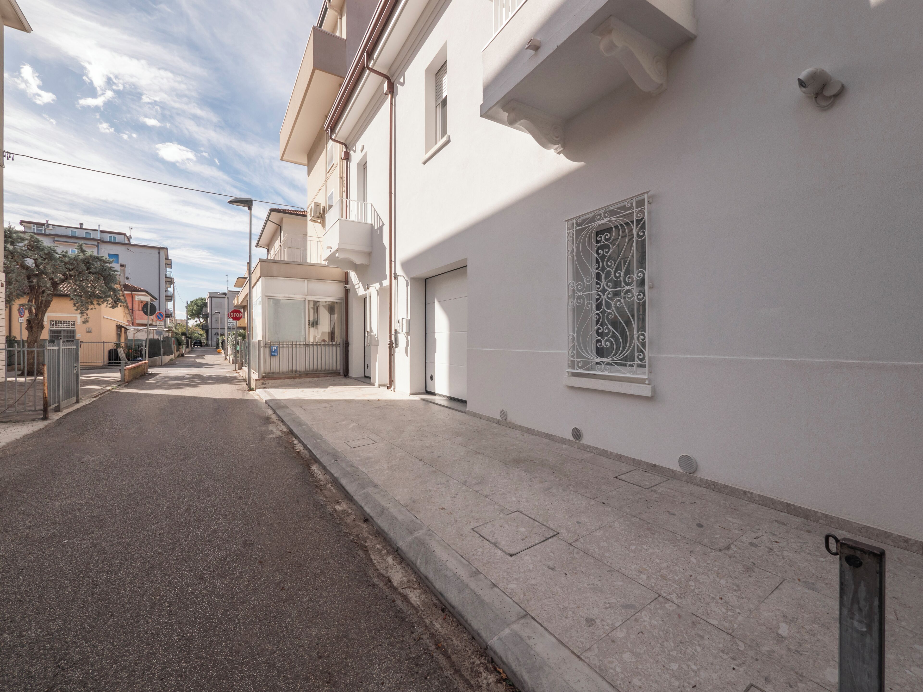 Una calle estrecha con un edificio blanco que tiene una reja metálica, un portón de garaje y un ventanal decorativo.