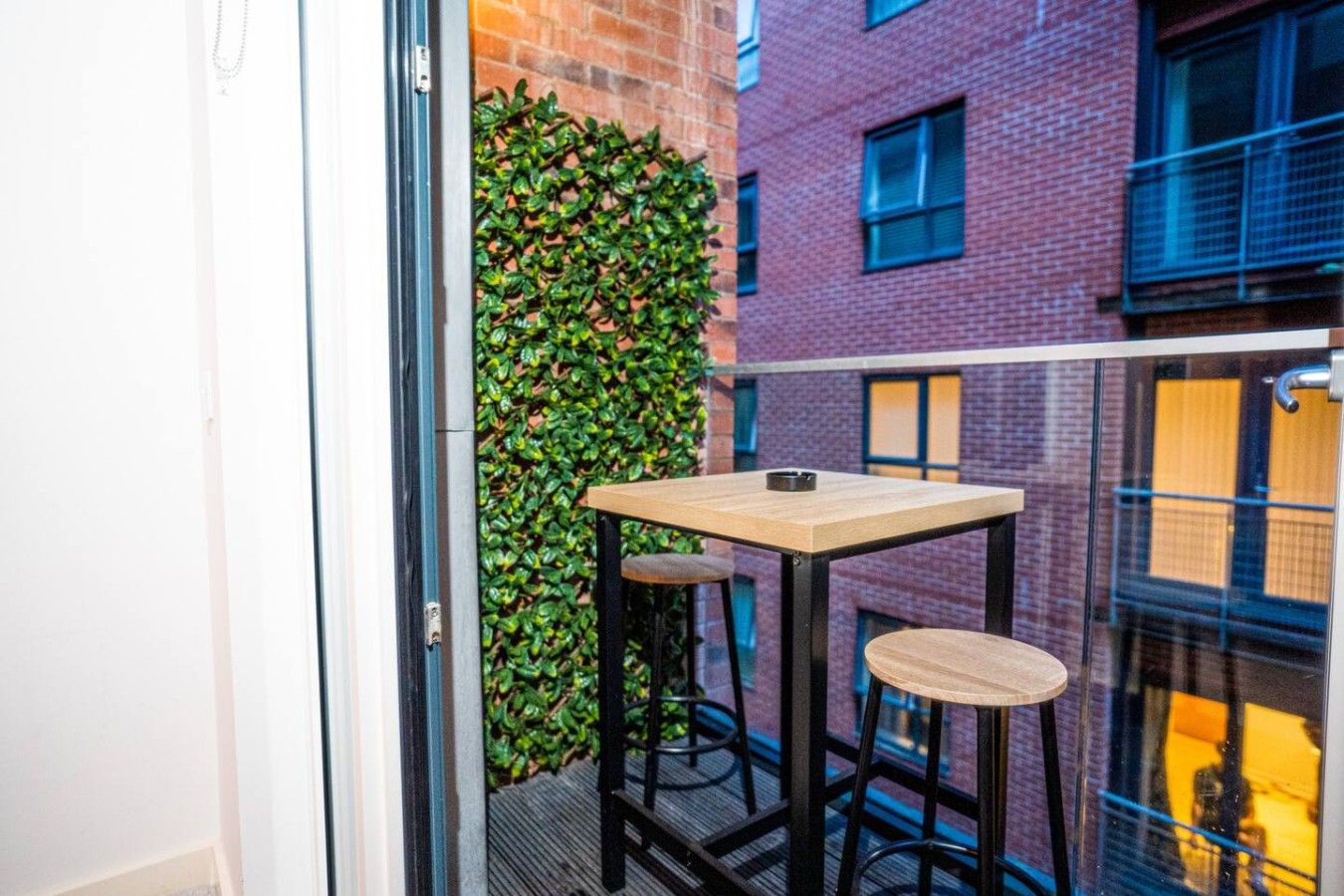 Un balcon avec une petite table et deux tabourets, une haie verte et, au fond, un bâtiment en briques.