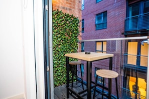 A balcony with a small table and two stools, a green hedge, and a brick building in the background. - City Centre Apt | Free Parking | Balcony | Airbed (Manchester)