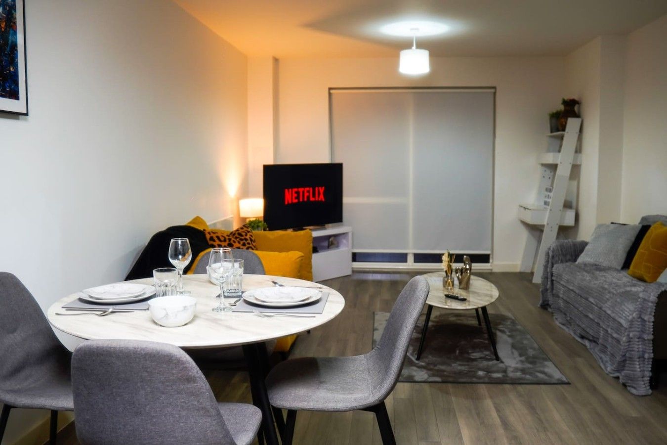 Een moderne woonkamer met een eettafel voor vier personen, een grijze bank, een televisie waarop 'NETFLIX' staat en een trap naar een hoger niveau.