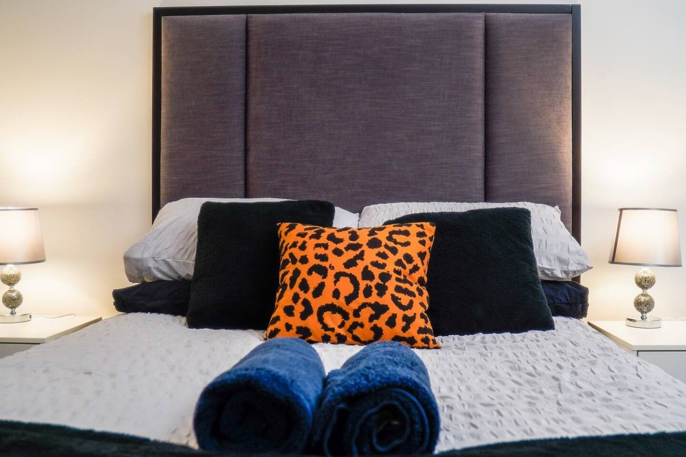 Un letto rifatto con un piumone bianco e nero, due asciugamani blu arrotolati e una varietà di cuscini, tra cui uno con una particolare fantasia leopardata arancione.