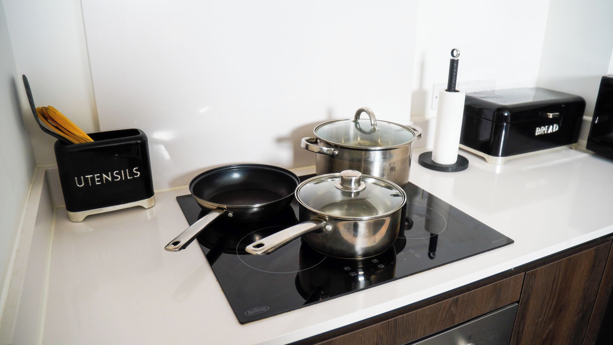 A kitchen counter with a black induction cooktop, two stainless steel pots, a frying pan, and a utensil holder labeled "UTENSILS".