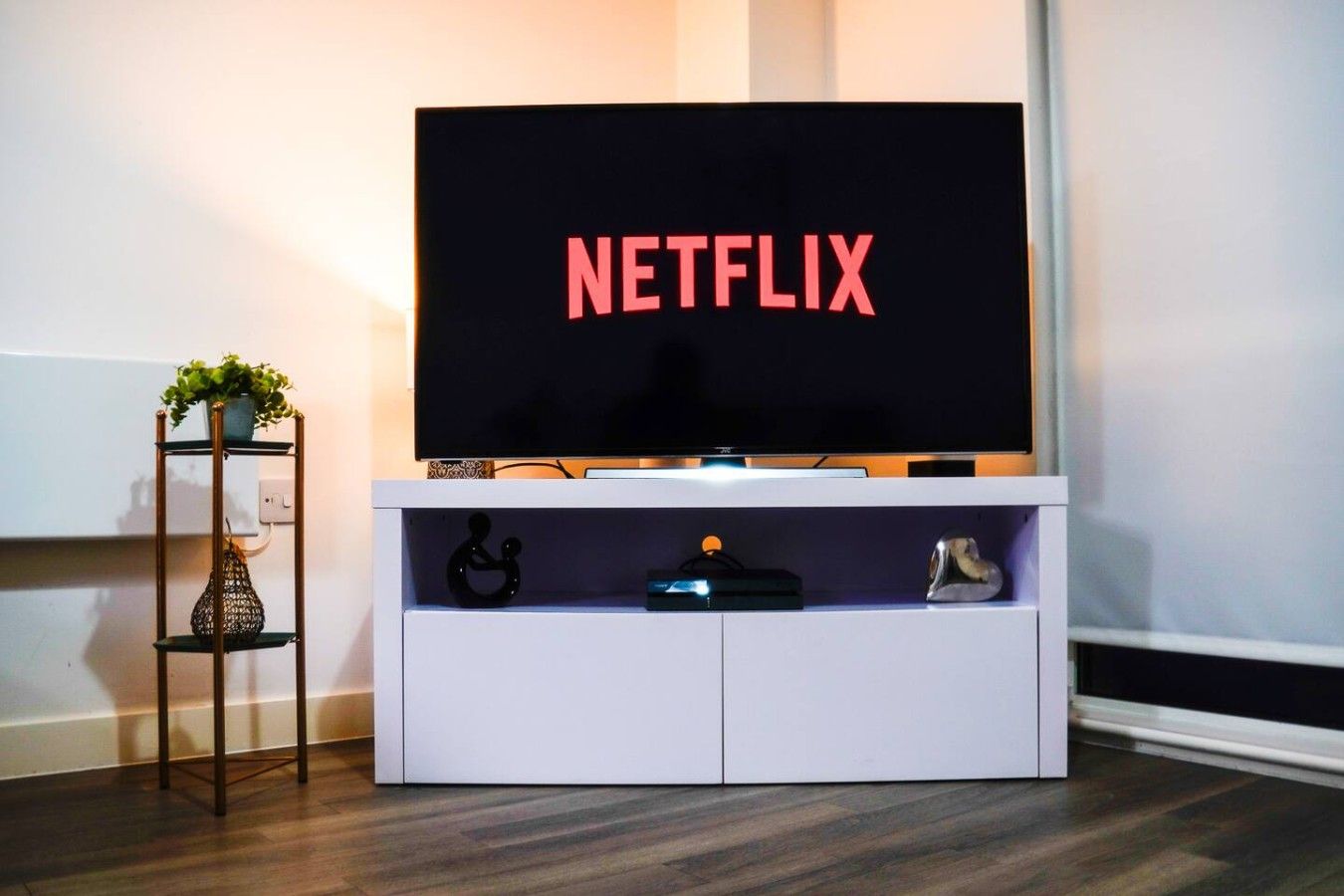 Un meuble de rangement blanc pour appareils électroniques, avec un téléviseur à écran plat affichant le mot « NETFLIX » en lettres rouges, une petite étagère ornée d’objets décoratifs et une plante posée sur un support métallique à côté.