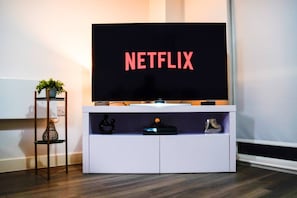 A white entertainment unit with a flat-screen TV displaying 'NETFLIX' in red letters, a small shelf with decorative items, and a plant on a metal stand beside it. - City Centre Apt | Free Parking | Balcony | Airbed (Manchester)