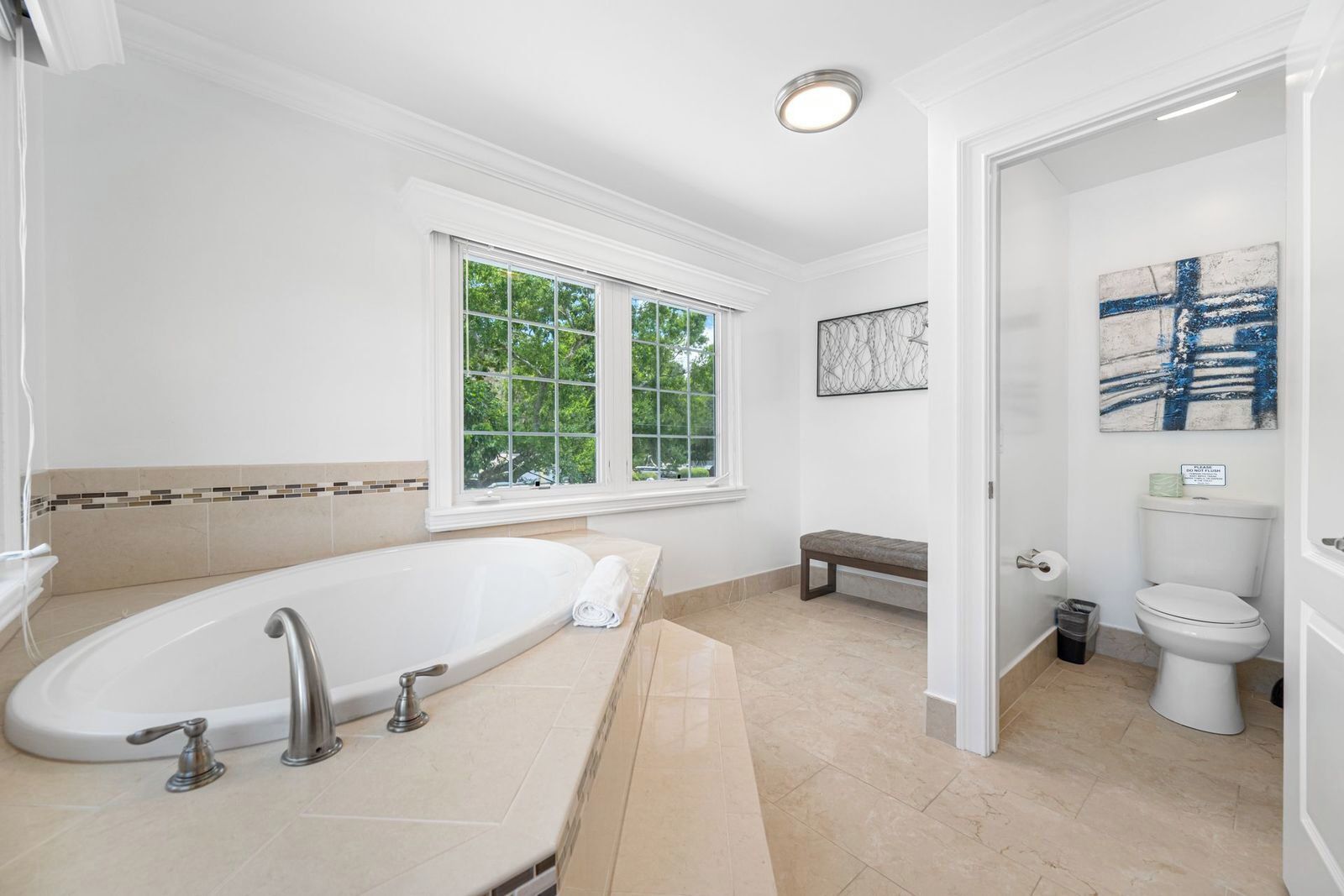 A modern bathroom with a freestanding bathtub, a bench, a toilet, and a window providing natural light.