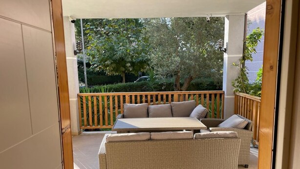 A covered outdoor seating area with a wicker sofa and two chairs, a wooden deck, and a view of trees and a car in the background.