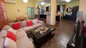 A living room with a sofa, coffee table, and a TV. There is a kitchen in the background with a dining area.
