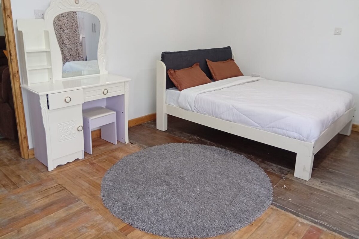 A bedroom with a white bed, a small white desk with a mirror, and a grey rug on the wooden floor.