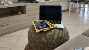 Ein modernes Wohnzimmer mit Flachbildfernseher, einem Laptop und einer Zeitschrift auf einem gewebten Hocker.
