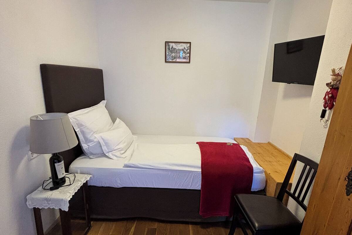 A small, neatly arranged bedroom with a single bed, a bedside table with a lamp, a chair, a TV mounted on the wall, and a picture frame.