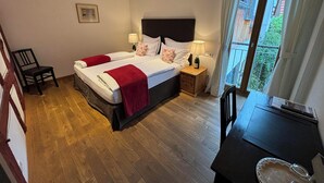 Habitación de hotel con dos camas, piso de madera, mesita de noche con lámpara, una silla y vista al balcón exterior.