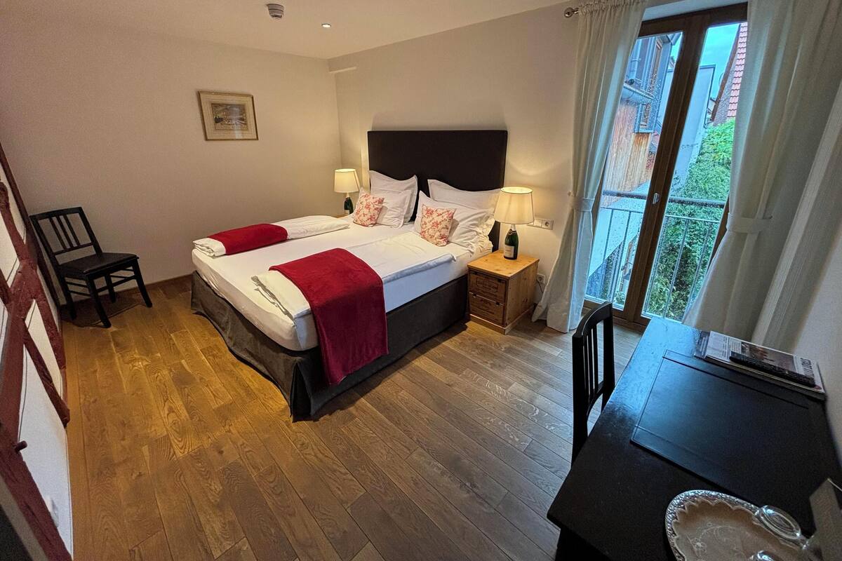 A hotel room with two beds, a wooden floor, a nightstand with a lamp, a chair, and a view of the outside balcony.