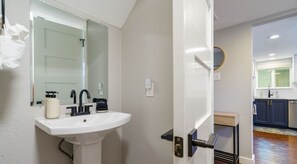 A bathroom with a white sink, black faucet, and a mirror. There is a door leading to a kitchen area with dark cabinets and a window. - Alexandria Dr xxxxxx (Austin)