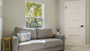 Ein stilvolles Wohnzimmer in neutralen Farbtönen mit einem grauen Sofa, einem kleinen Holzschemel und einem runden Hocker. Eine weiße Tür und ein Fenster mit Blick auf Bäume sind zu sehen.