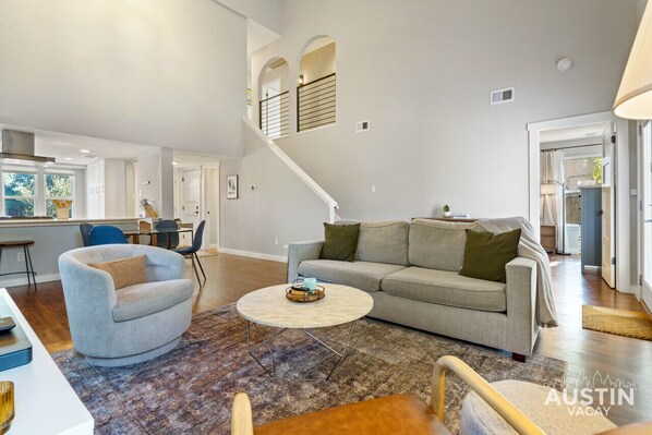 A spacious living area with a grey sofa, a round coffee table, and a grey armchair. A staircase with a white railing leads to an upper level. - Alexandria Dr xxxxxx (Austin)