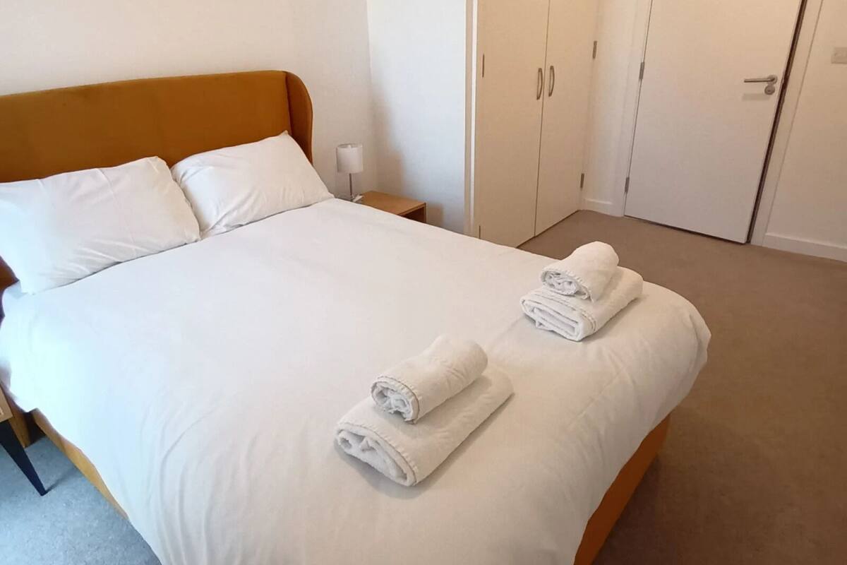 Una cama bien hecha con sábanas blancas y dos toallas enrolladas, un cabecero de madera, una mesita de noche con una lámpara y un armario de dos puertas.