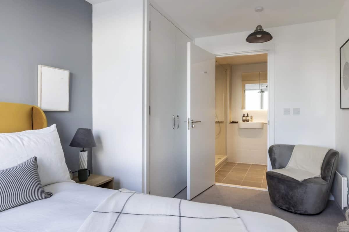 Un dormitorio moderno con cama, mesita de noche, lámpara y un sillón gris.