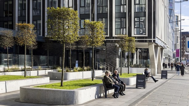 Un edificio moderno con facciata in vetro e un'area pedonale con panchine e alberi.