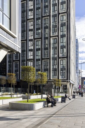 Un bâtiment moderne doté d’une façade en verre et d’un espace piétonnier agrémenté de bancs et d’arbres.
