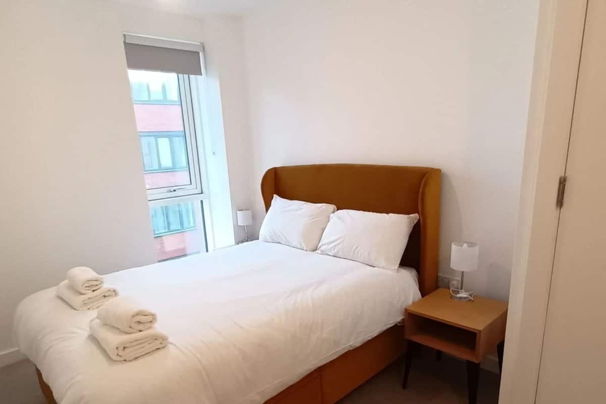 Un dormitorio con cama, dos almohadas, una mesita de noche, una lámpara y una ventana.