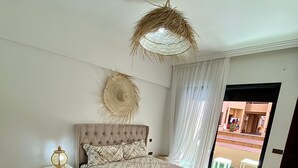 Un dormitorio con cabecera tapizada, cama con colcha rosa, lámpara de techo tejida, tapiz de pared tejido y vista a un patio exterior a través de una puerta de cristal.