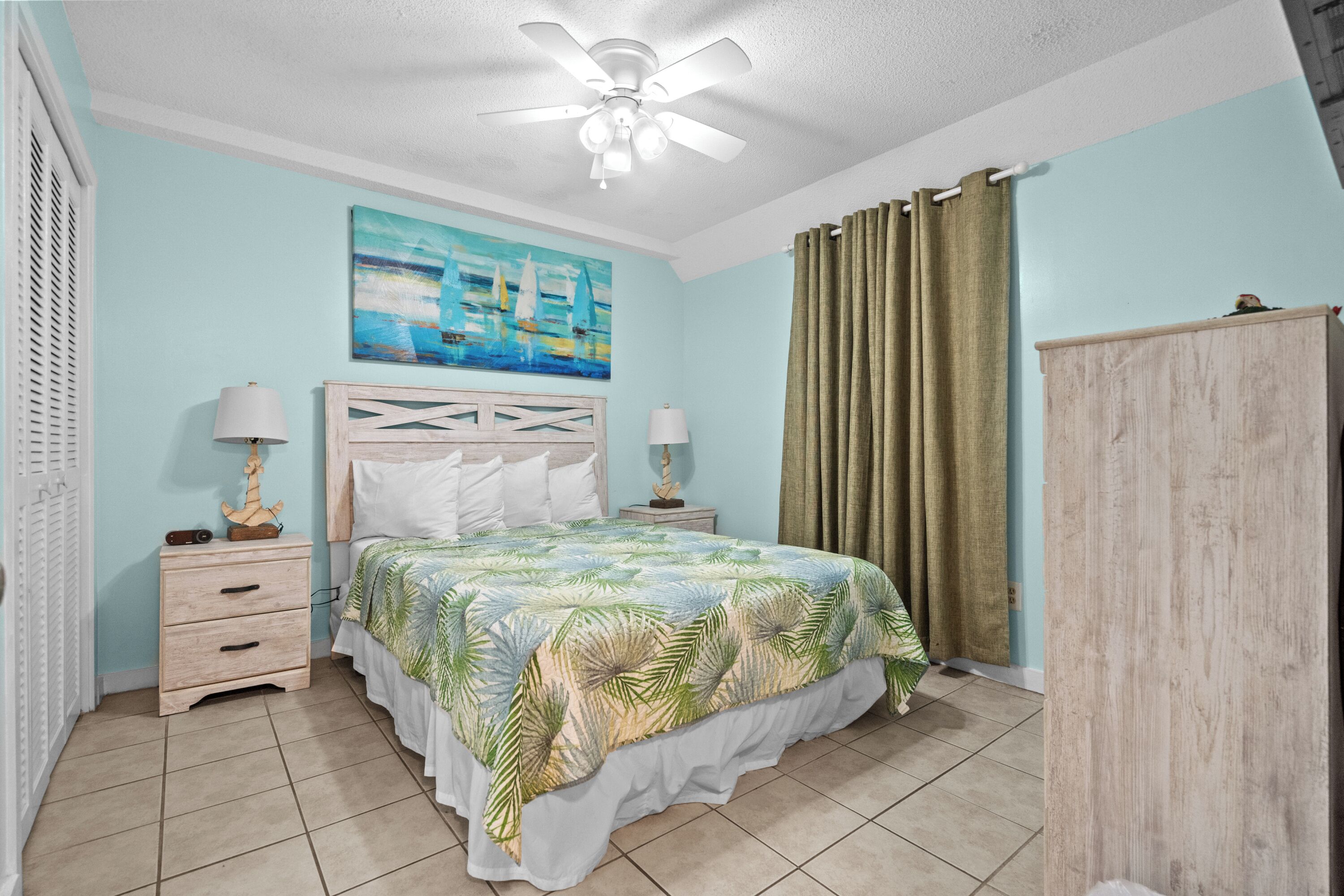 A bedroom with a bed, two bedside tables, a nightstand, a ceiling fan, a painting, and a wardrobe.