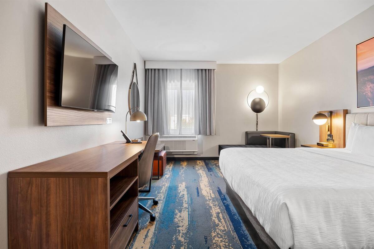 Een hotelkamer met een groot bed, een bureau met stoel, een aan de muur bevestigde televisie, een bureaulamp en een raam met gordijnen.