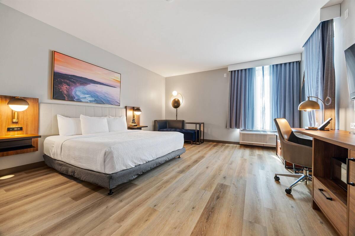 Een hotelkamer met een bed, bureau, stoel en een groot raam met gordijnen.