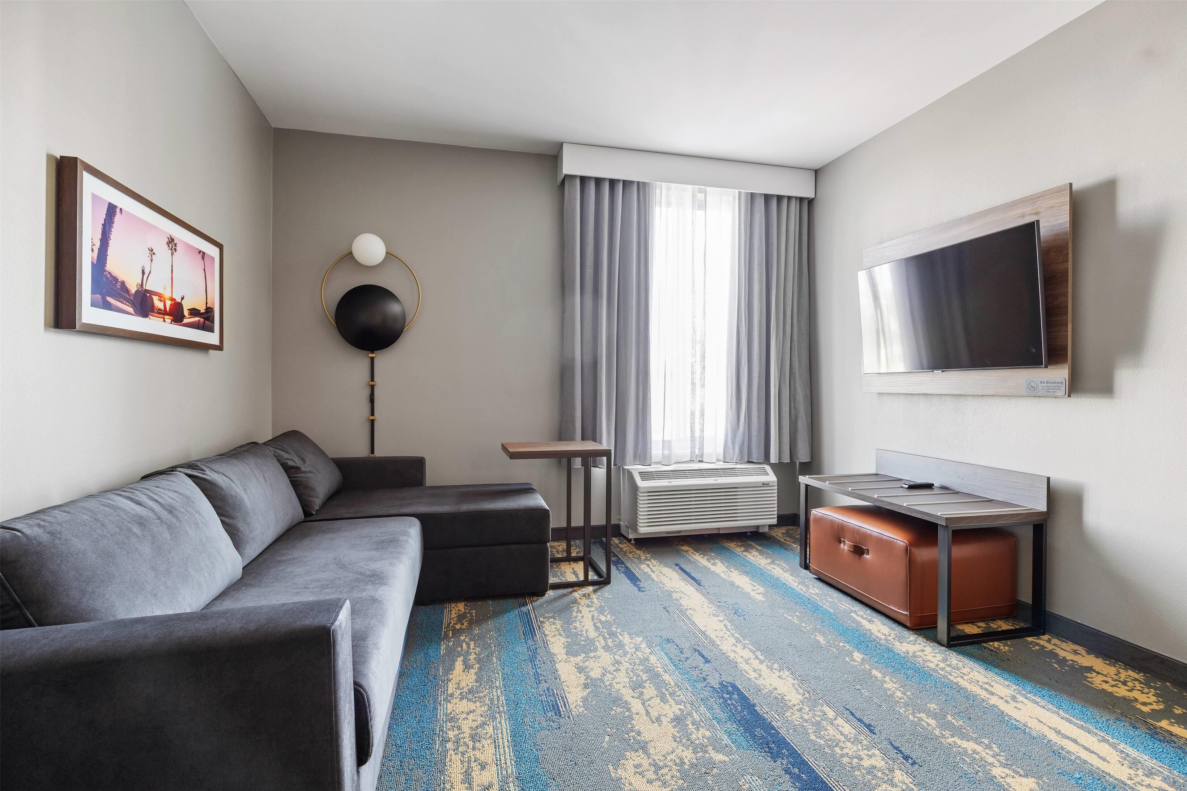 A modern hotel room with a grey sofa, a flat-screen TV, a small table, a lamp, and a suitcase.