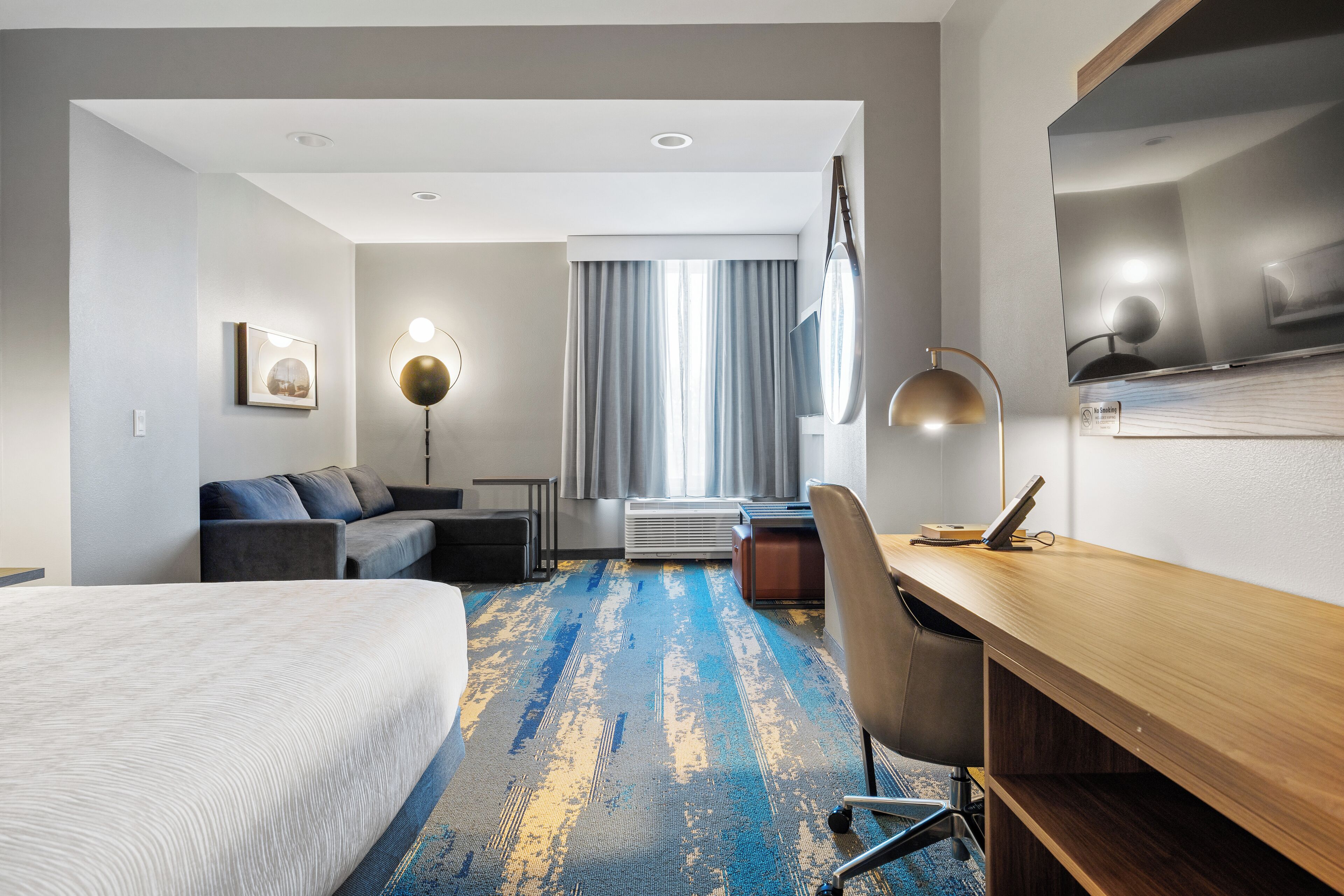 A hotel room with a large bed, a desk, a chair, a sofa, a floor lamp, and a TV.