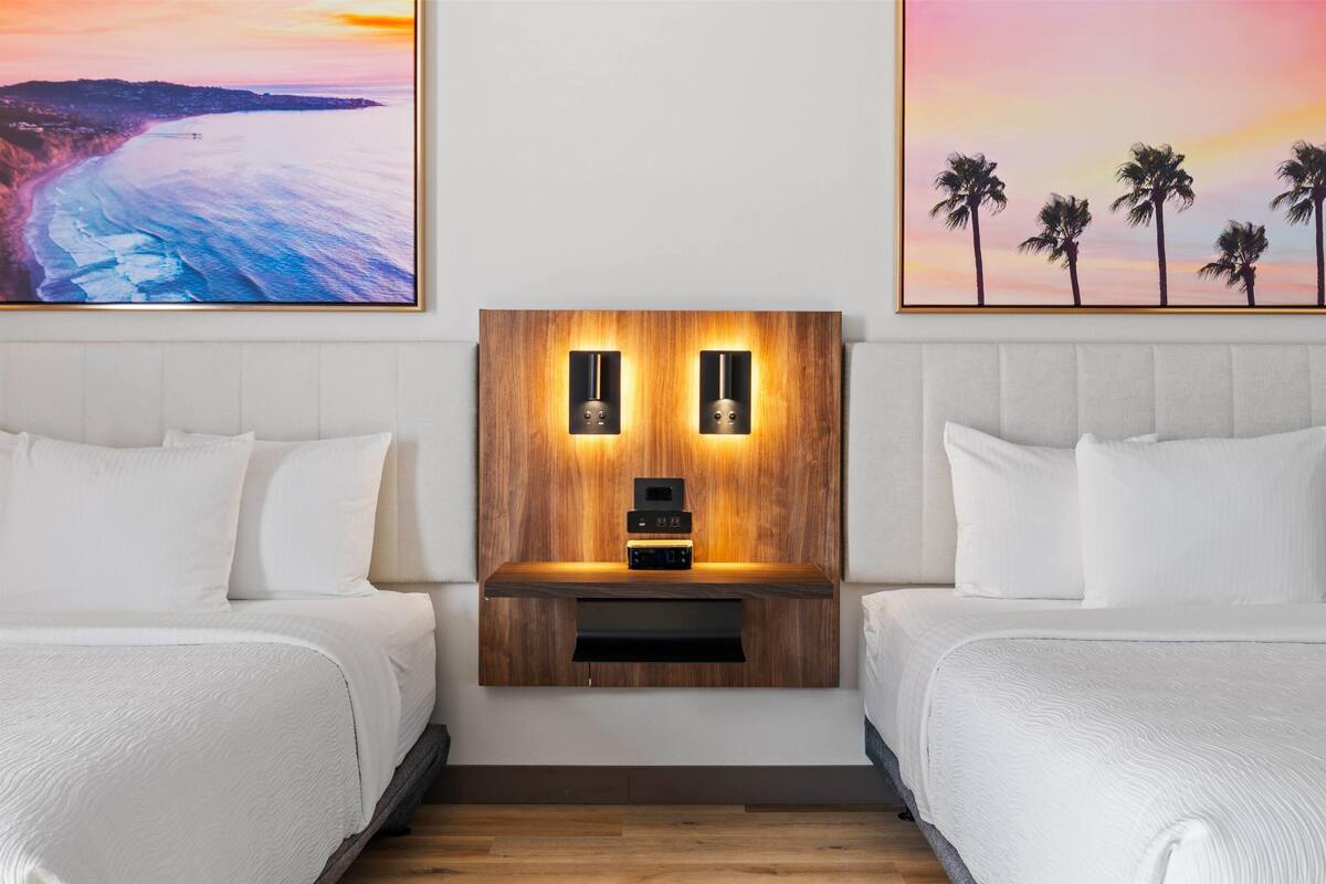 Een hotelkamer met twee bedden, een houten hoofdeinde met ingebouwde verlichting en kunstwerken die een strand en palmbomen voorstellen.