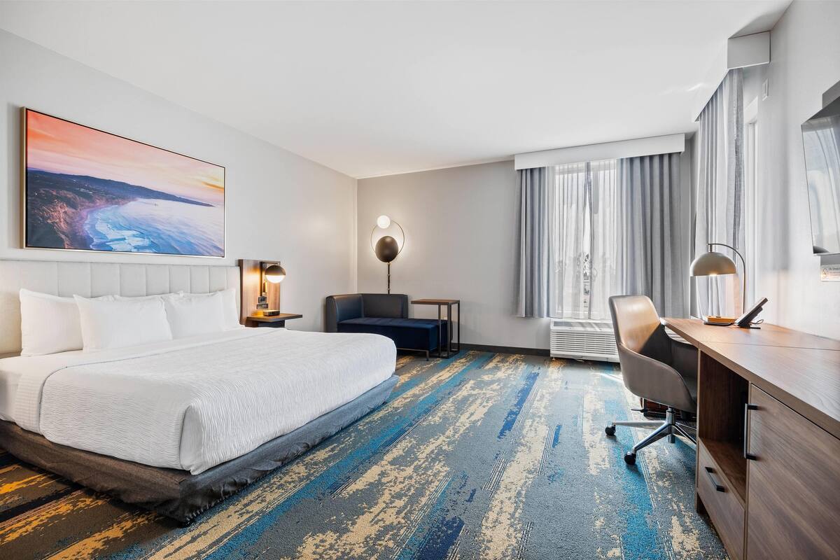 Hotelkamer met een groot bed, een bureau, een stoel, bureaulamp, vloerlamp en een schilderij van een zonsondergang boven de oceaan.