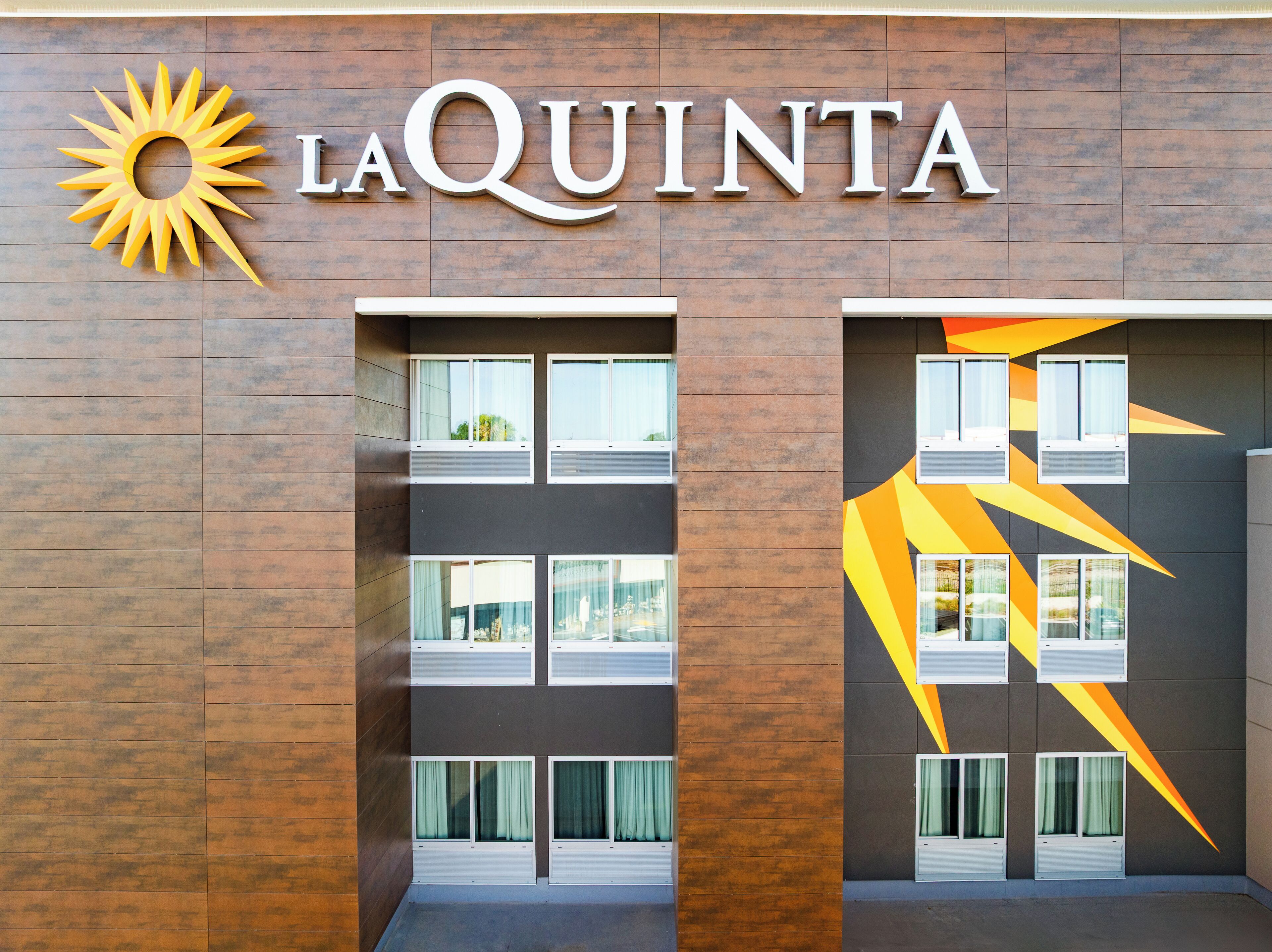 Fachada de hotel con el nombre "LA QUINTA" y un logo de sol. Se observan varias ventanas, algunas con cortinas, y un diseño geométrico a la derecha.