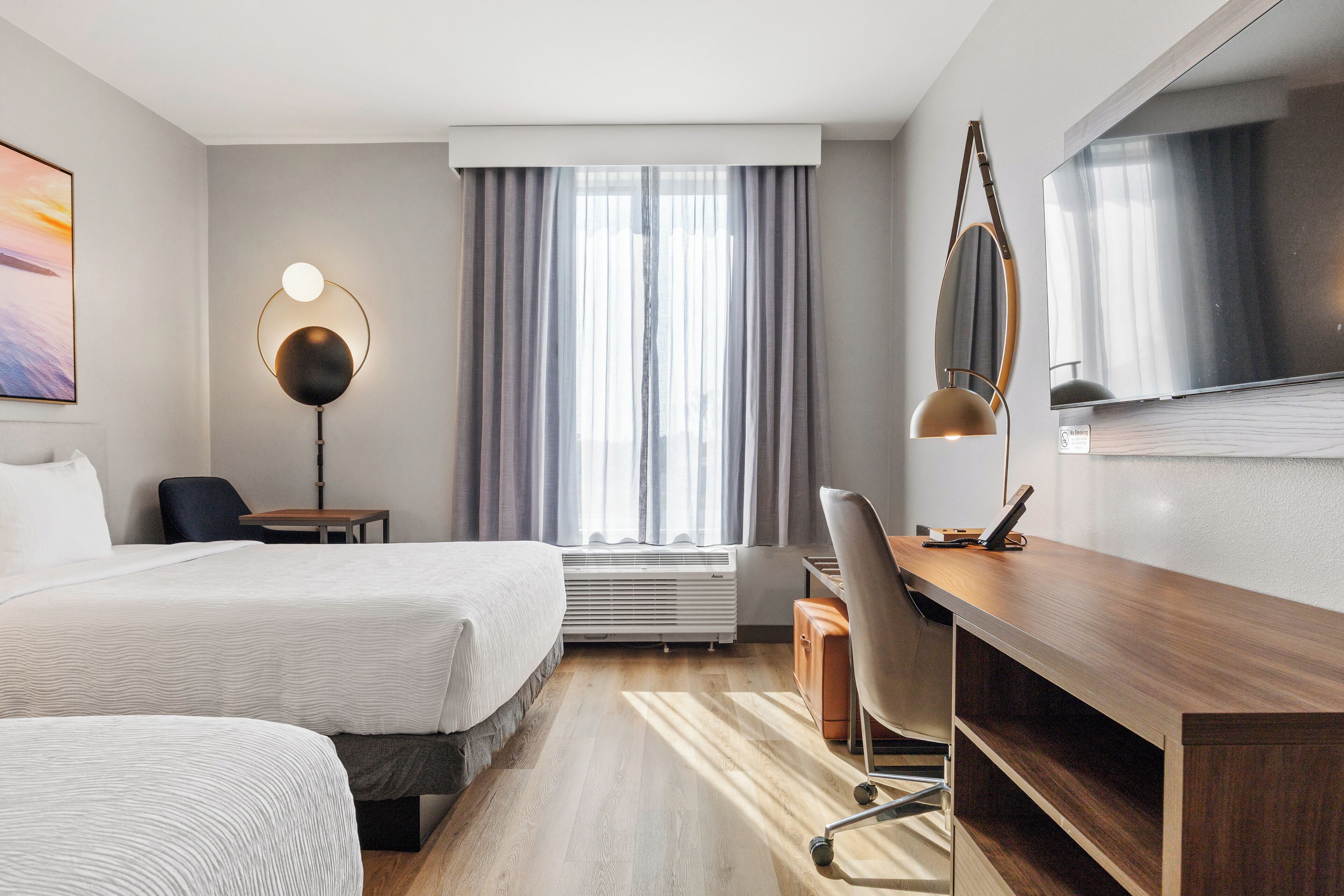 A hotel room with two beds, a desk, a chair, a TV, a lamp, and a window with curtains.