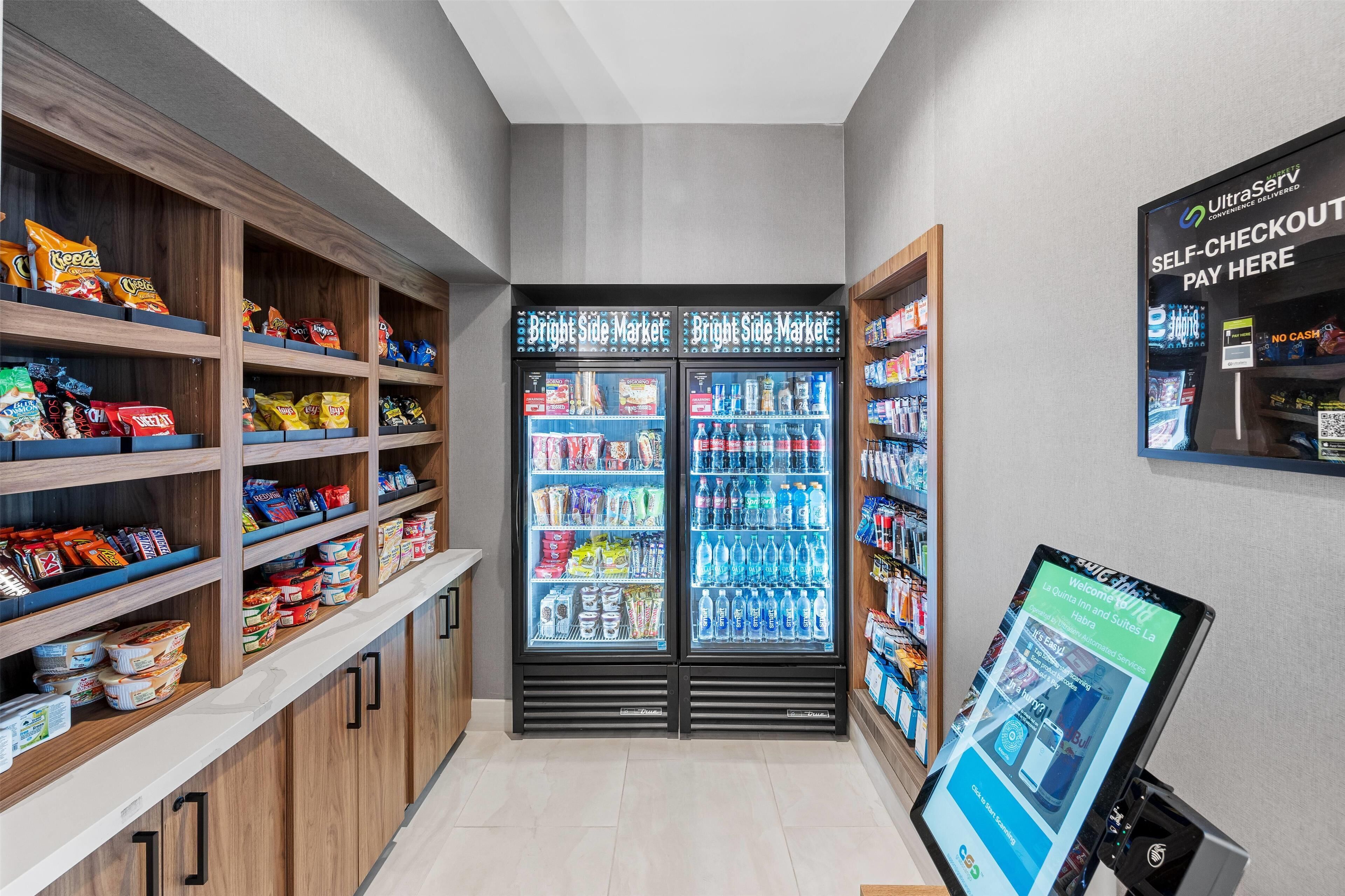 A self-checkout area with a digital screen, snack shelves, and refrigerated beverage cases.