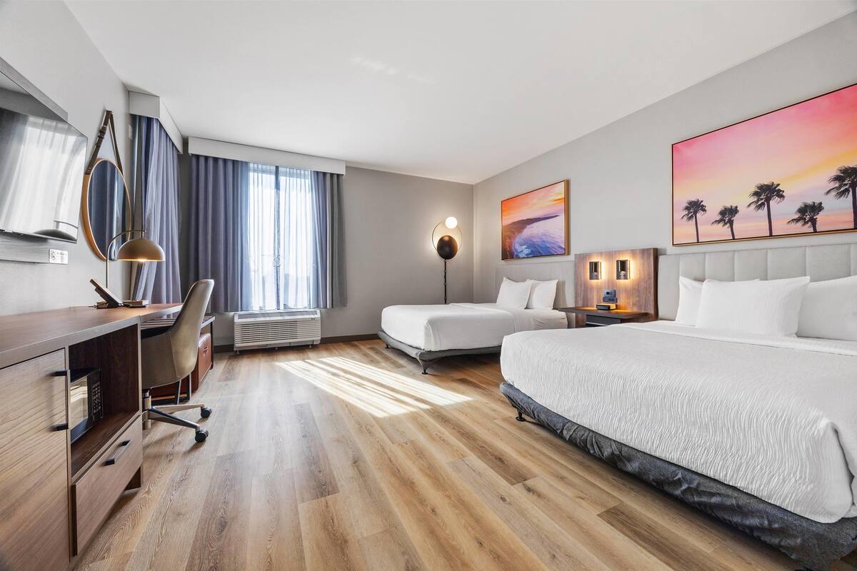Een moderne hotelkamer met een houten vloer, een bureau met stoel, een bed met wit beddengoed, een nachtkastje met lamp, en een groot schilderij van een zonsondergang.